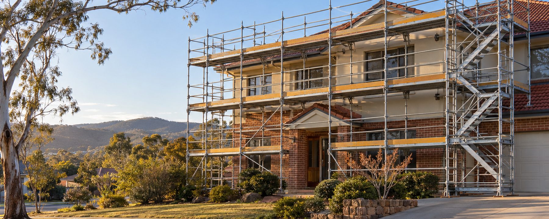A double-storey brick and tile house in Canberra with a four-lift modular steel scaffold across the front, a stair tower at one end, a ghost gum beside it and hills in the background